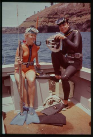 Shot of Valerie Taylor with spear rod and Ron Taylor with underwater film equipment both suited in dive gear