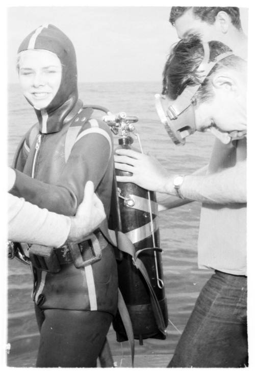 Black and white shot of Cathy Trout in scuba diving gear being assisted by two people