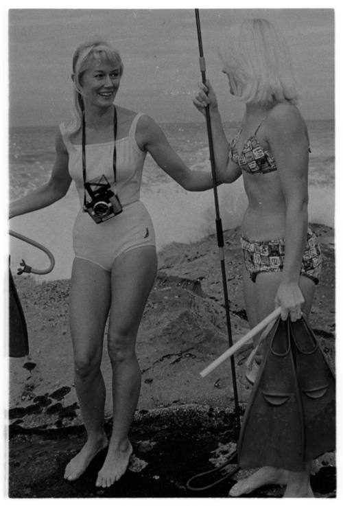 Black and white shot of two people in swimsuits holding snorkelling equipment and spear rod standing on beach shore