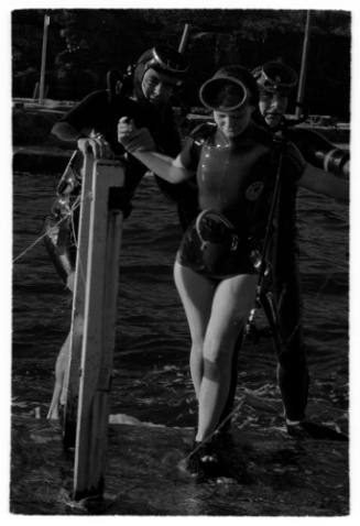 Black and white shot of two divers in full gear assisting a student diver walking through shallow water