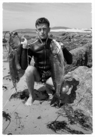 Black and white shot of diver sitting on rocky shore holding up two caught fish