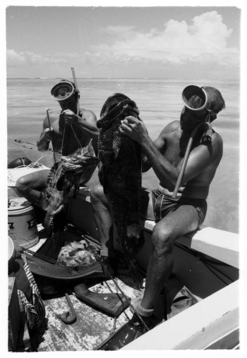 Black and white topside shot of two divers each holding a caught fish sitting on a dinghy at sea