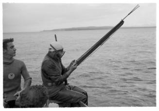 Black and white shot of three divers sitting on a dinghy at sea, with one sitting holding a spearfishing gun on the edge at the stern