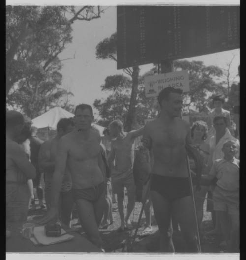 Shot of person Ron Taylor holding two caught fish standing by crowd at a weighing in area