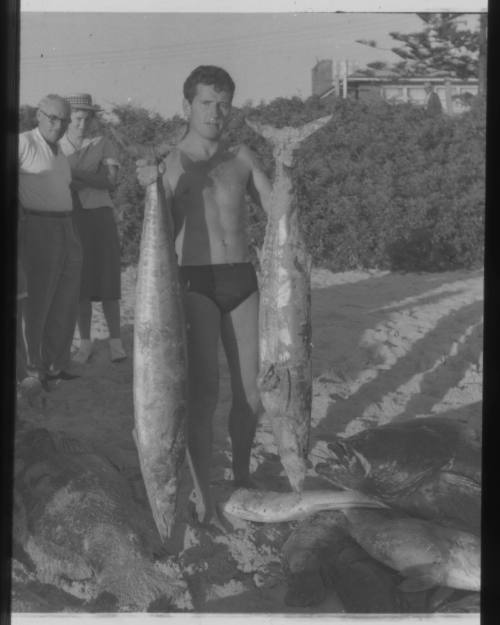 Shot of person holding two caught fish nearly their own height amongst more of their catch on the floor next to them on the beach