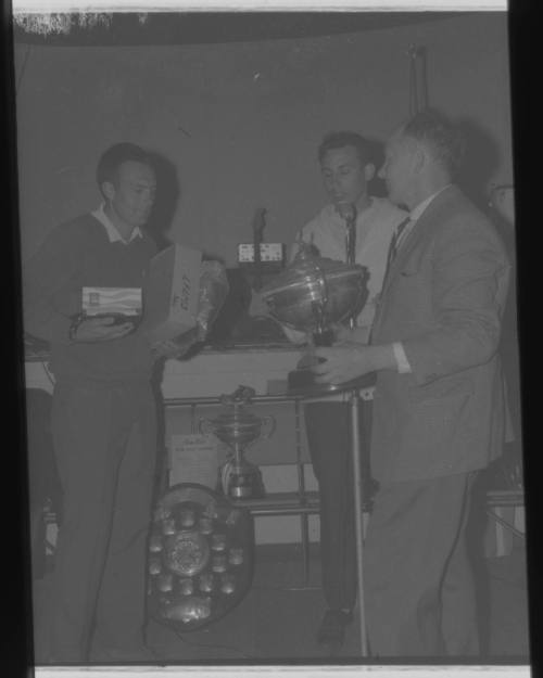 Shot of two people holding trophy and boxes with another person and more trophies in background