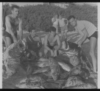 Shot of four people holding and surrounding their catch on the floor at the beach, with two children posing in background