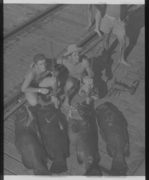 Shot of two people holding spearguns crouched by their caught fish by tracks on ground