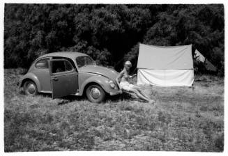 Black and white shot of Volkswagen parked grass with person sitting on hood and tent pitched in background
