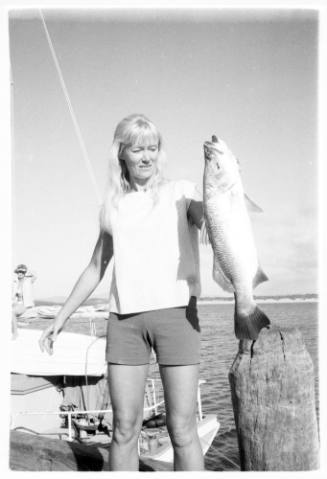 Black and white shot of person holding fish by gill standing on the side of a wharf, with dinghy in background