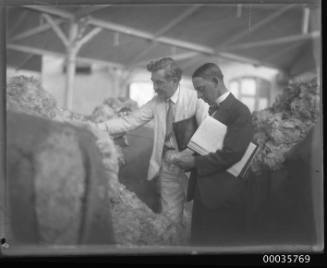 Interior view of two men in suits with folders handling wool from open bales.