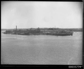View of Cockatoo Island from west Balmain showing wharf, warehouses and a chimney.