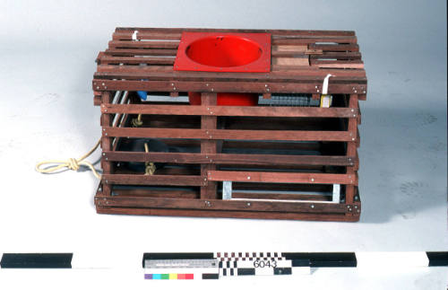 WOODEN FRAME, RED PLASTIC NECK, RECTANGULAR BASE, SLANTED SIDES, SMALLER RECTANGULAR TOP, A SMALL SECTION OF THE LID NEAR THE NECK LIFTS OUT TO REVEAL A SMALL METAL CAGE, YELLOW NYLON  ROPE ATTACHED