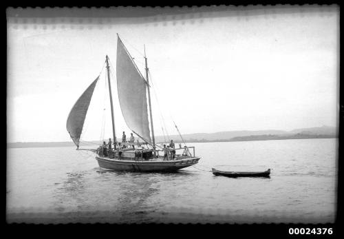 Crew on the deck of a ketch under sail towing a long dinghy