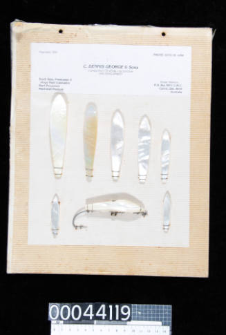 An album page with eight pieces of trimmed and polished pearl shell, one attached to a metal fishing hook, attached with string 
