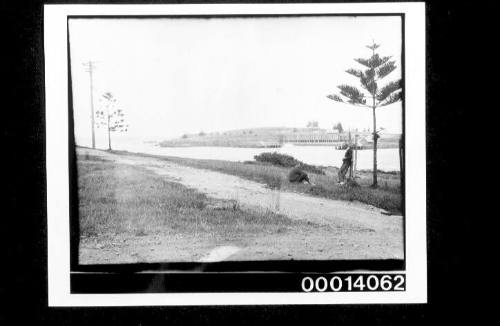 Two men standing on a grassy foreshore, wharf across the water in the distance