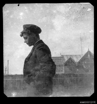 Negative of a man in a jacket and hat, standing side on, with a wooden fence visible in the background