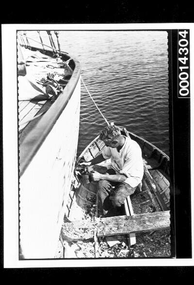 A young man in a dinghy removes paint from the hull of a yacht