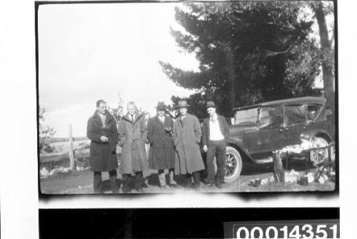 Group of men wearing coats standing beside a car, water in the distance