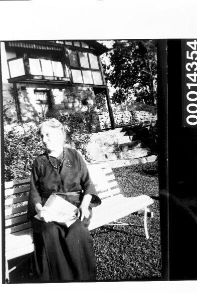 An elderly woman seated in a garden outside a home