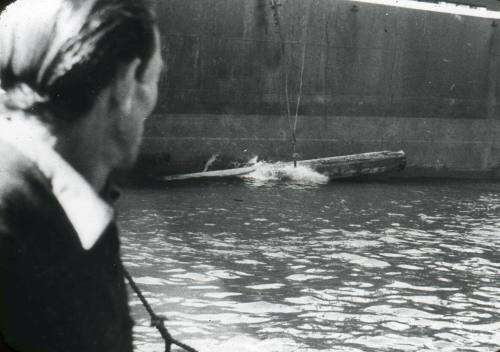 Slide depicting a large wooden log tied with cable falling into water alongside a ship