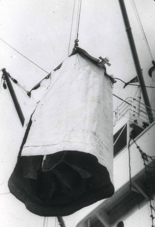 Slide depicting an item shrouded in canvas being transported by crane on or off a ship