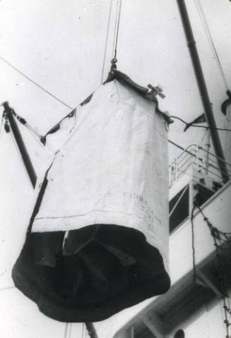 Slide depicting an item shrouded in canvas being transported by crane on or off a ship