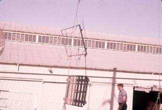 Slide depicting a wooden pallet hanging vertically from cables in front of a warehouse