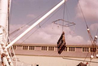 Slide depicting an pallet hanging vertically from a square frame suspended from a crane