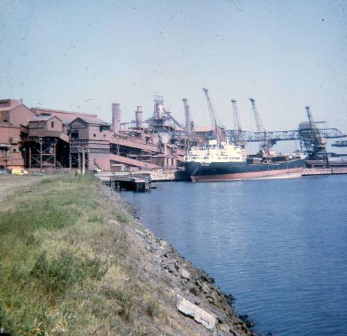 Slide looking across water to a cargo ship alongside an industrial wharf