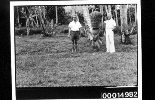 Two men stand at either side of a large carved Tahitian idol