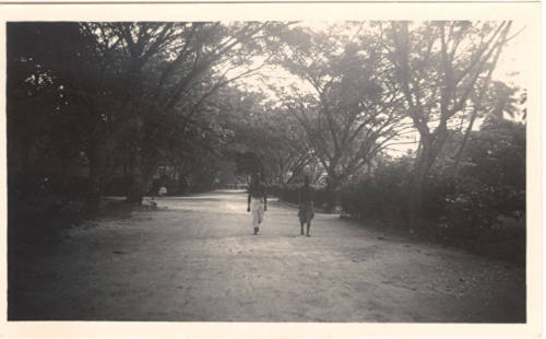 Photograph depicting two men walking down a dirt road in a tropical landscape