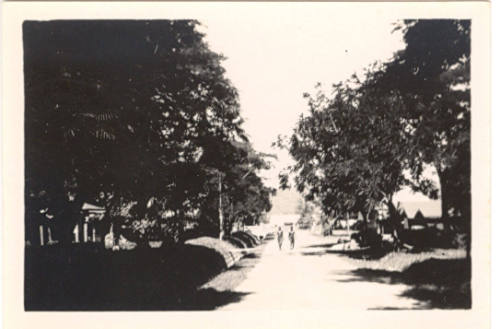 Photograph depicting two men walking down a dirt road in a tropical landscape