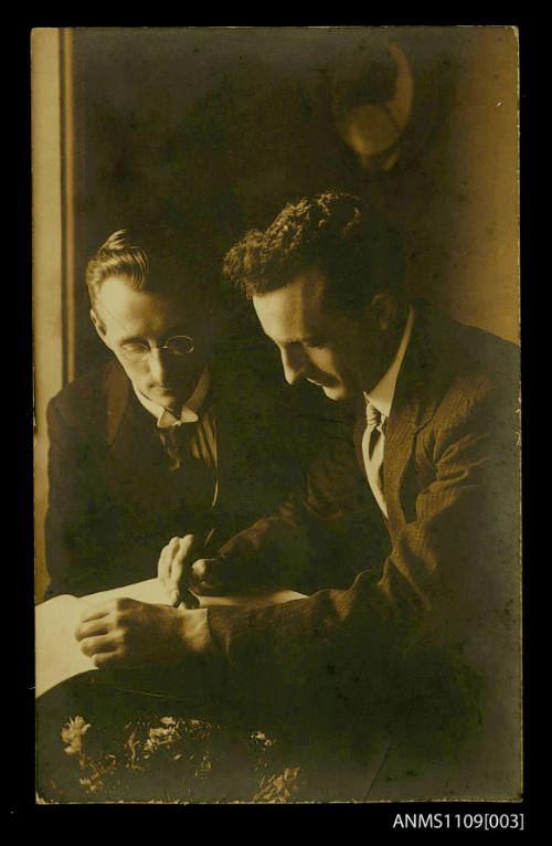 Photographic postcard of two men wearing suits and looking at a page