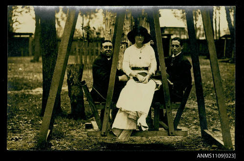 Photographic postcard of a two young men and a woman on a wooden swing