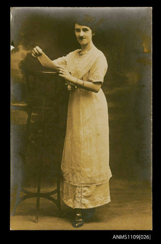 Photographic postcard of a woman dressed in white leaning on a high table