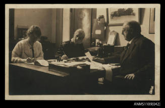 Photographic postcard of a man and two women seated at partners desk