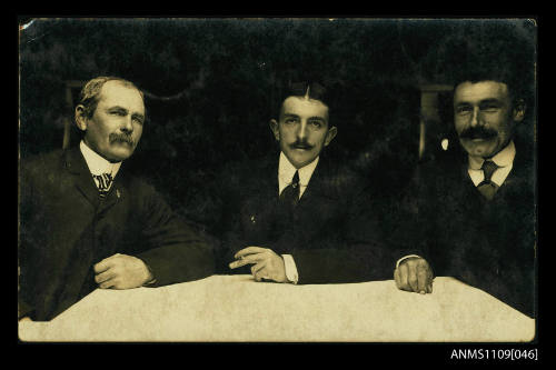 Photographic postcard of three men with moustaches sitting around a table