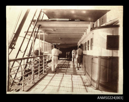 Deck of unknown ship, left is railing and rigging with the water just visible, on the left in the distant are two people seated against railing