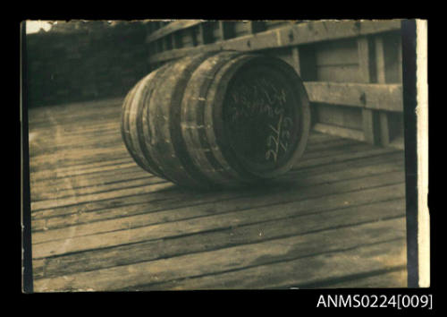 Photograph of wooden barrel on wooden wharf with wooden wall on right with barrel on side