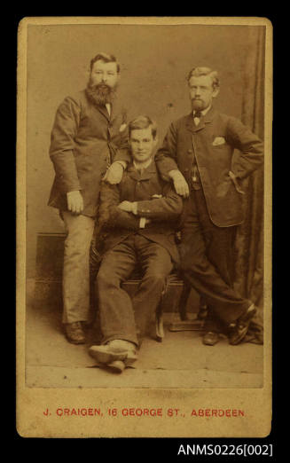 Group photograph of three men. Two men stand behind seated male in front. On left, adult male, dark jacket, light trousers, dark shoes, large dark moustache and beard