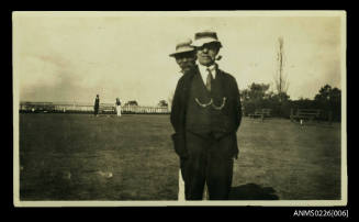 Two older men facing camera, on bowling green. Centre, man in dark suit, waistcoat with two chains to pockets. Hands in trouser pockets. Soft hat. Pipe in mouth