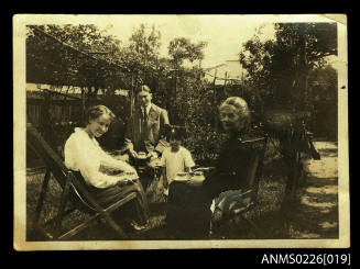 Two women, man, seated in garden with female child, on left, woman, white blouse, dark skirt, seated on deck chair