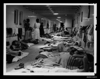 Enclosed deck of passenger ship with people lying in rows and standing looking out of portholes